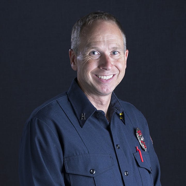 A man in a uniform with badges, smiling against a dark background.