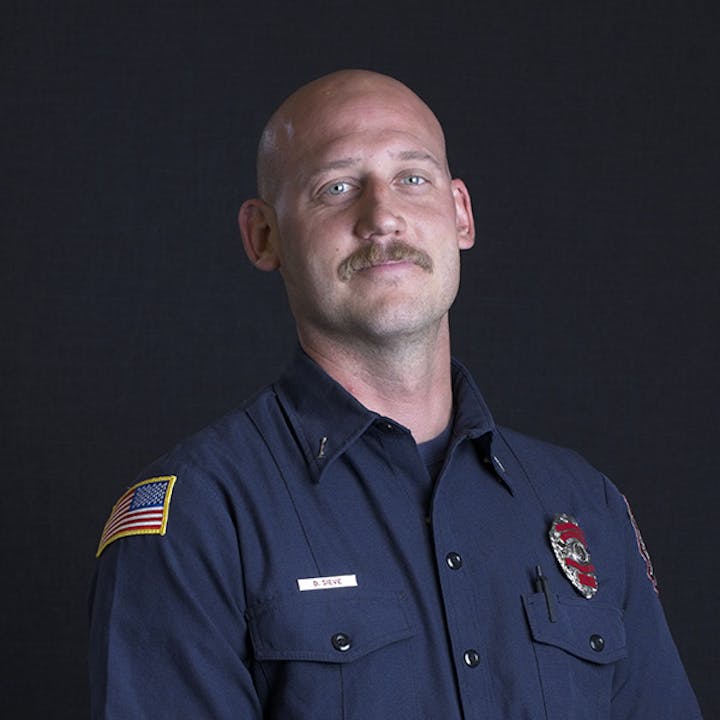 A person in a dark uniform with a badge and flag patch, against a plain background.
