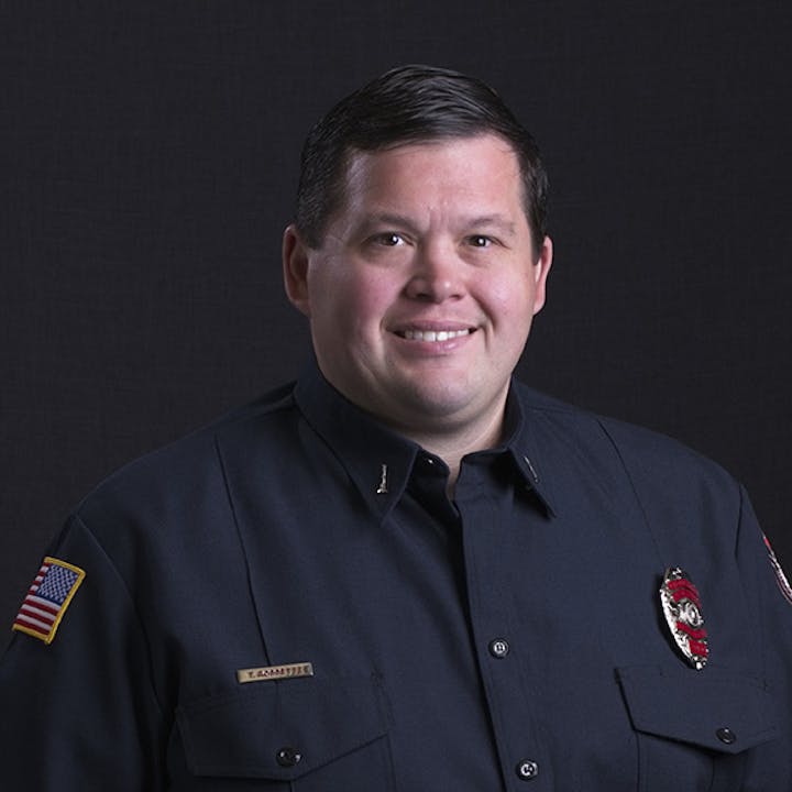 A man in a dark uniform, with an American flag patch and a badge, smiling against a dark background.