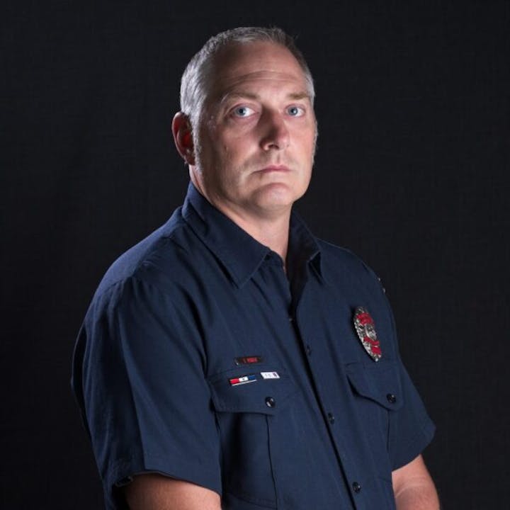 A person in a navy blue uniform with badges on the chest, against a dark background.