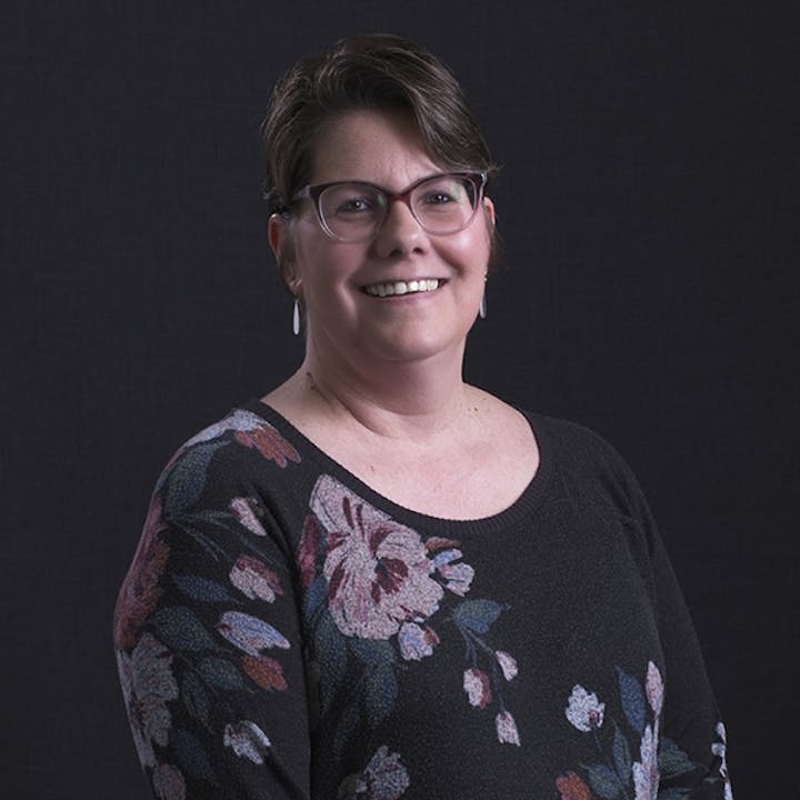 A person smiling, wearing glasses and a floral-patterned sweater against a dark background.