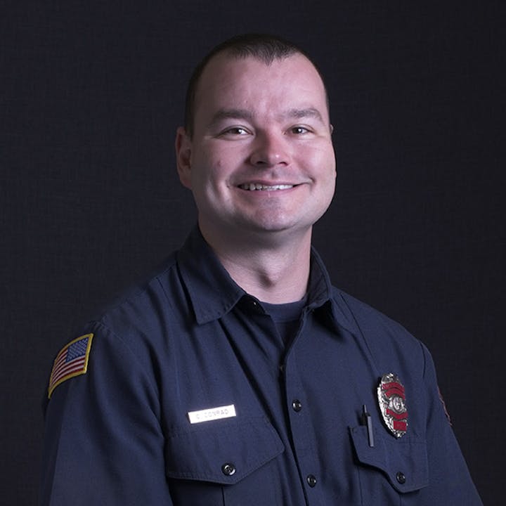 A person in a firefighter uniform with an American flag patch, smiling against a dark background.