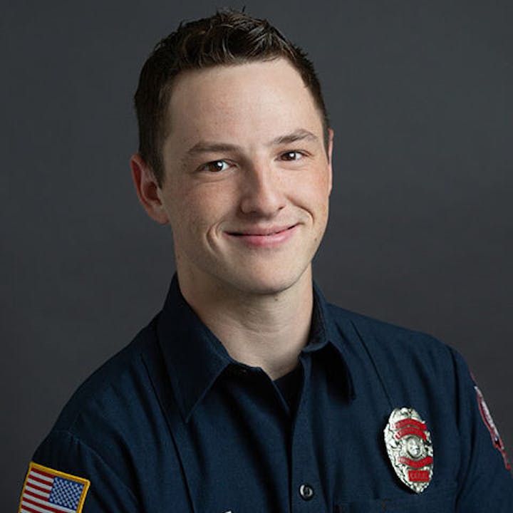 A person in a firefighter uniform smiling at the camera against a gray background.