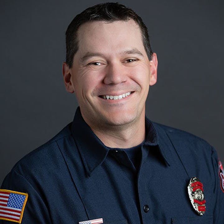 A smiling person wearing a dark uniform with badges and an American flag patch.
