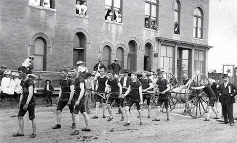 A group of men in uniforms pulling a cart in a parade, with onlookers in front of a brick building.