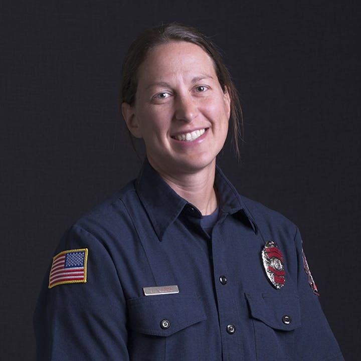 A person wearing a dark uniform with American flag and badge, smiling against a dark background.