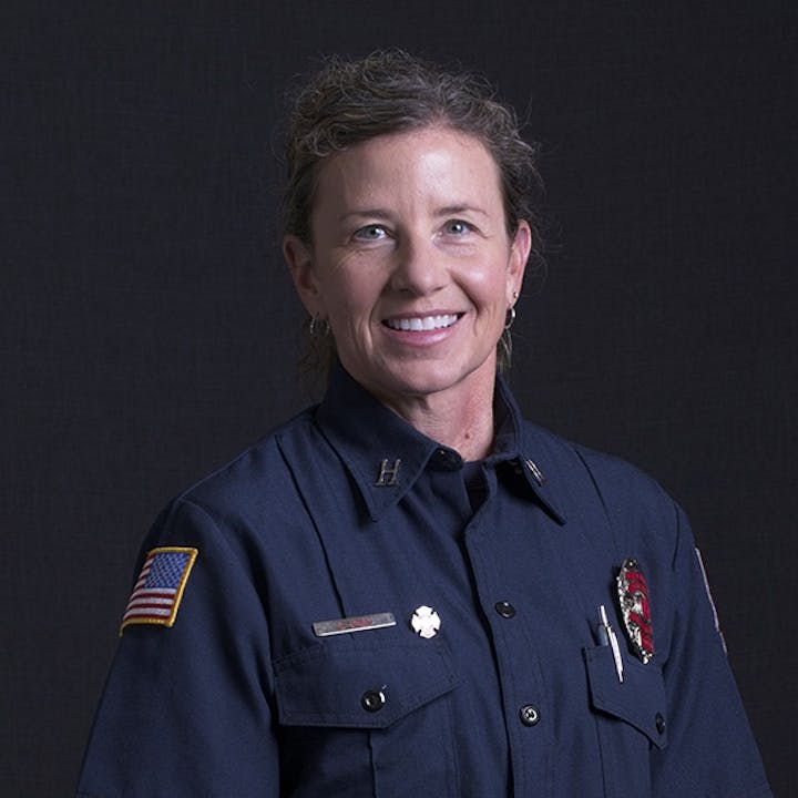 A person in a firefighter uniform with badges and an American flag patch, smiling, against a dark background.
