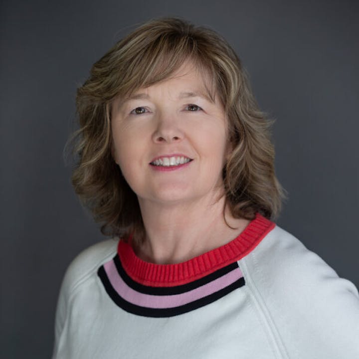 A person with shoulder-length hair, wearing a sweater with red and pink stripes on the collar, smiles at the camera.