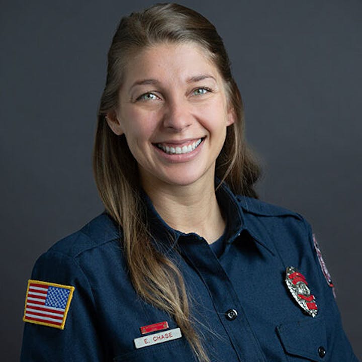 A person in a uniform with badges and an American flag patch, smiling against a dark background.