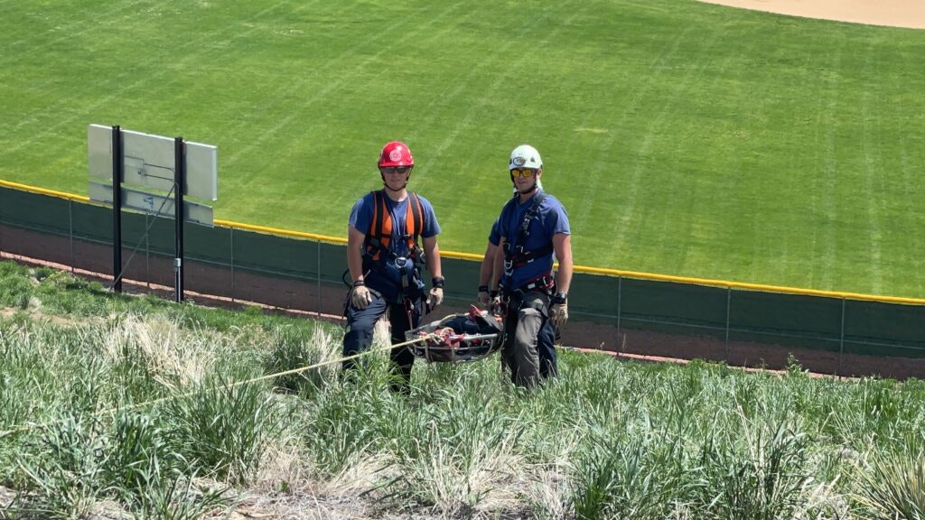 Two workers in safety gear stand on a grassy slope with a sports field in the background.