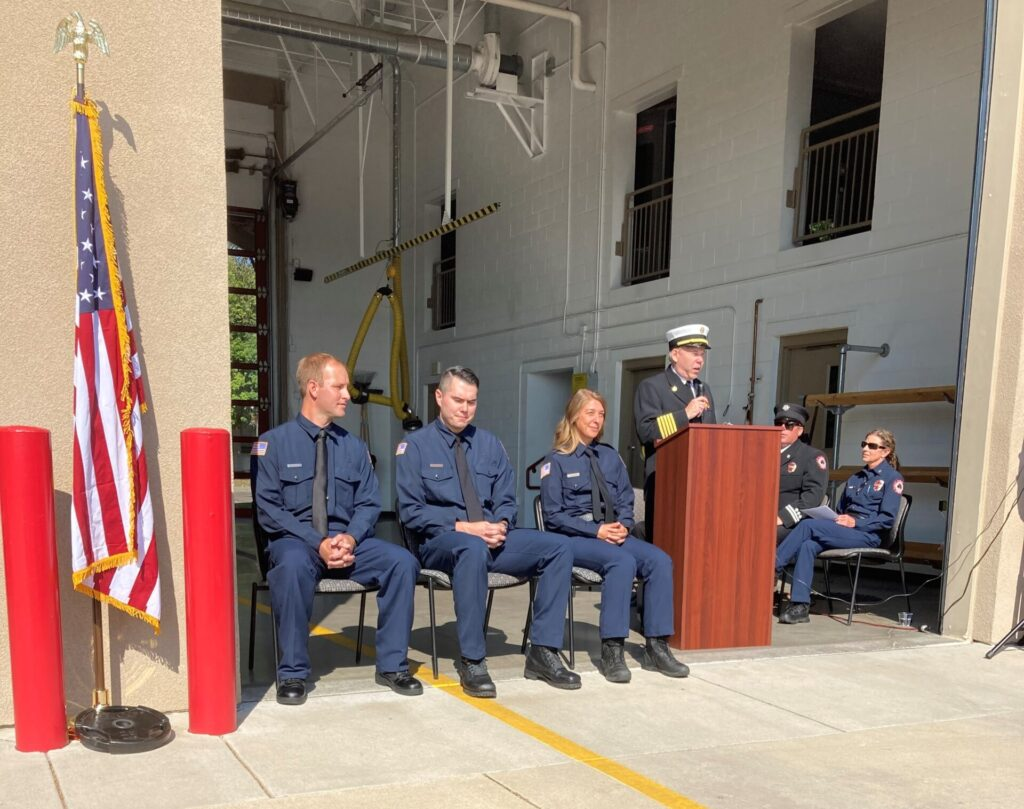 A group of uniformed firefighters seated, with one standing at a podium speaking, near an American flag, in an open garage setting.