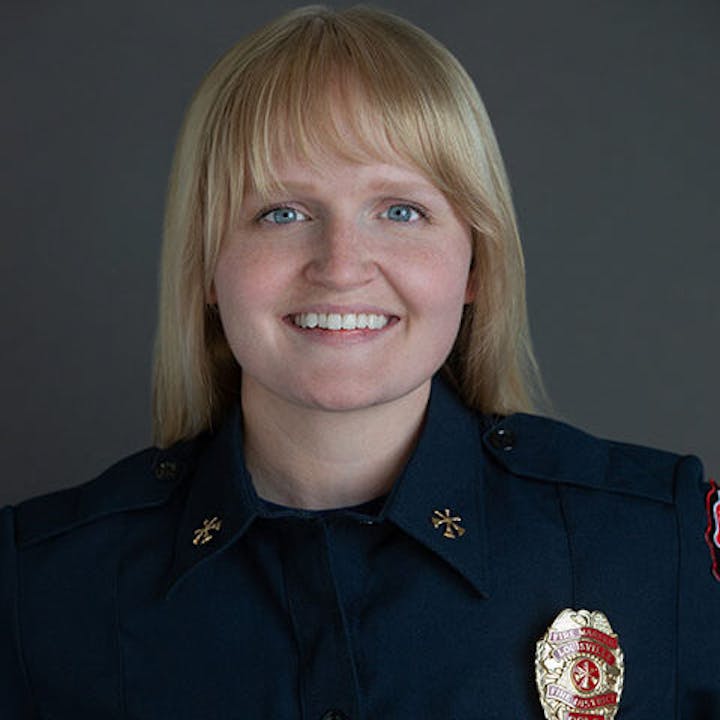 A person smiling, wearing a dark uniform with a fire department badge.