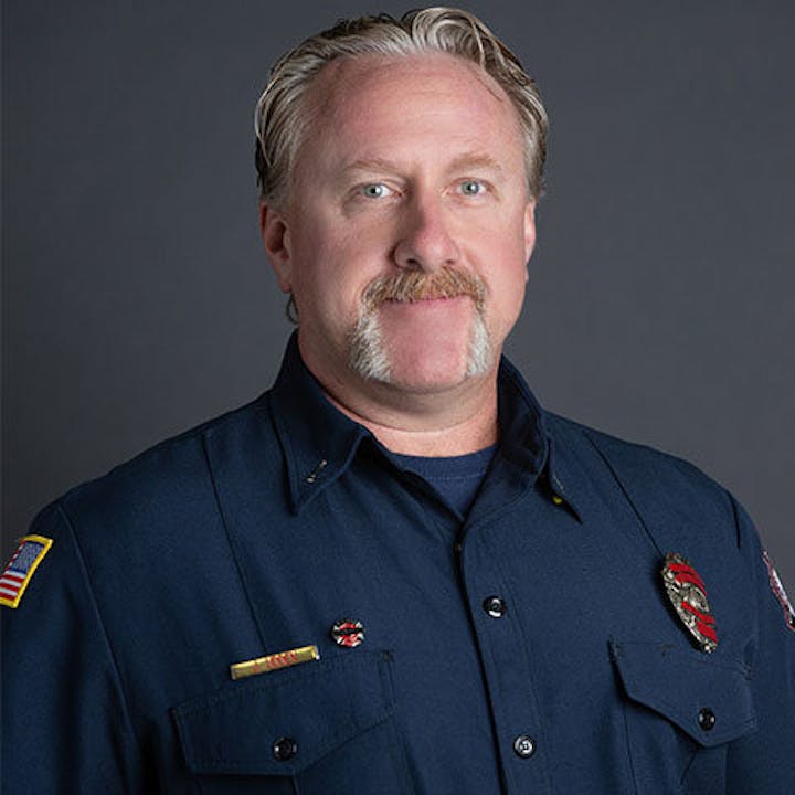 A person in a dark uniform with badges, likely a firefighter or public safety officer, against a gray background.