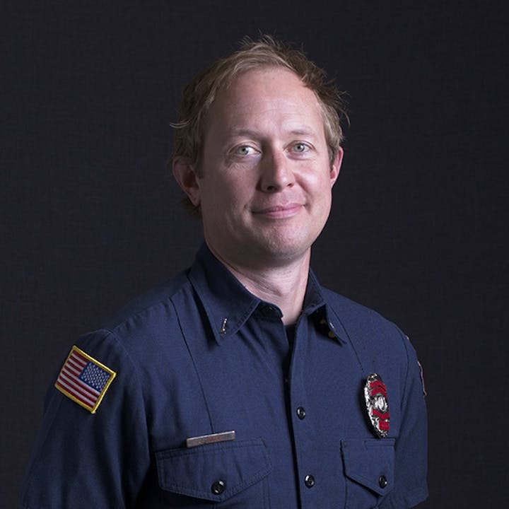 A person in a dark uniform with an American flag patch and a badge, against a dark background.