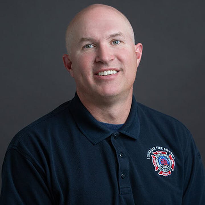 A man wearing a navy blue collared shirt with a fire department logo, smiling against a dark background.