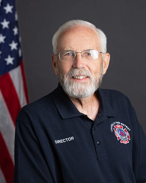 An older man with glasses and a beard wearing a fire district shirt labeled "Director" stands in front of a U.S. flag.