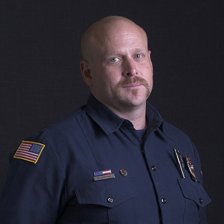 A man in a dark uniform with badges, an American flag patch on the sleeve, and a mustache, against a dark background.
