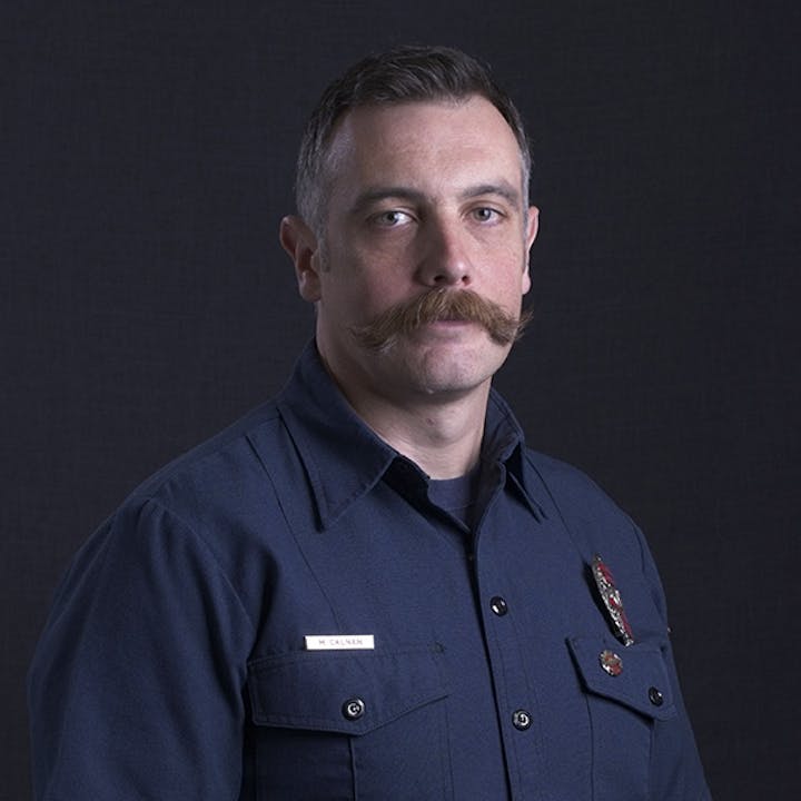 A man with a mustache in a dark uniform, wearing a badge, against a dark background.