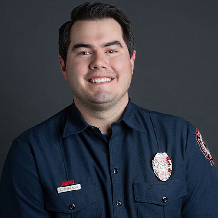 A person in a blue uniform with a badge, smiling, in front of a dark background.
