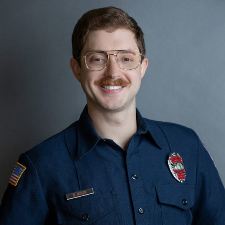 A person in a blue uniform with glasses and a mustache, smiling.