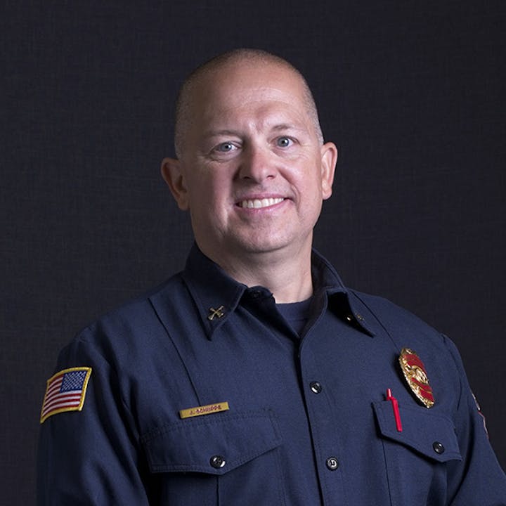 A person in a dark uniform with a badge and an American flag patch on the sleeve, smiling against a plain background.