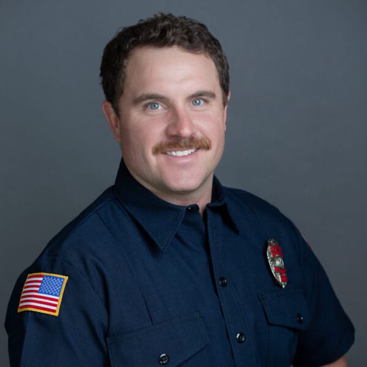 A smiling man in a navy blue uniform with an American flag patch and a badge, posing for a portrait against a gray background.
