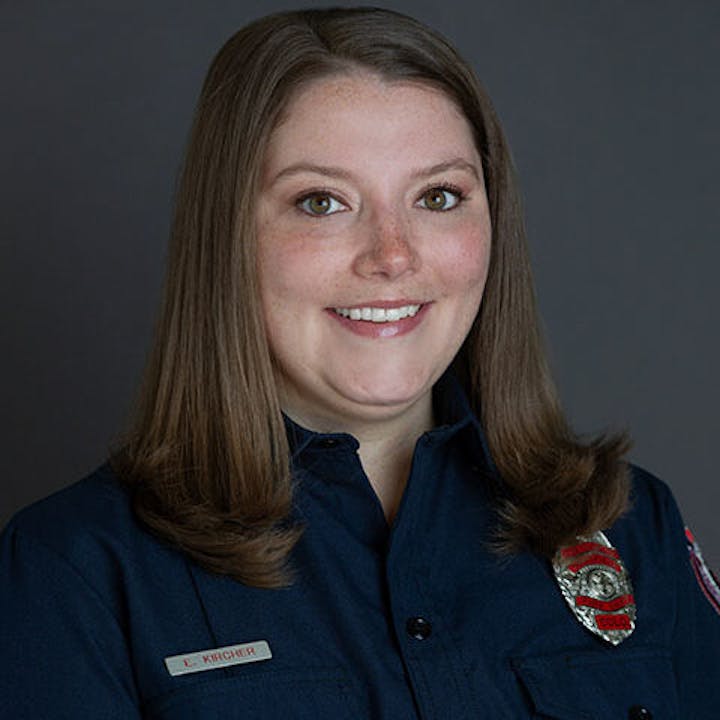 A person in a dark uniform shirt with a badge, smiling in front of a plain background.