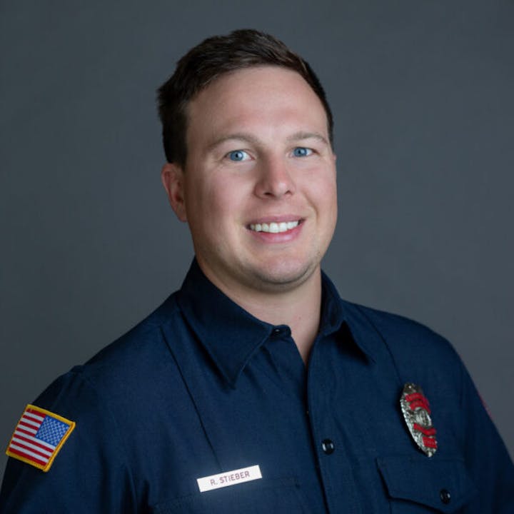 A person in a firefighter uniform with a name tag, badge, and flag patch, smiling against a neutral background.