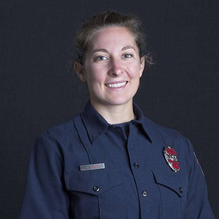 Smiling person in a uniform with a badge on the chest, against a dark background.