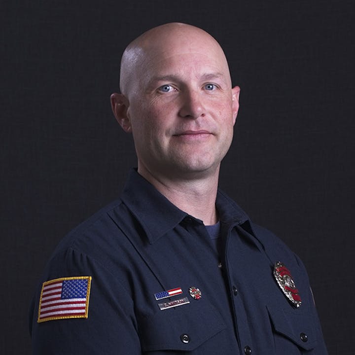 A person in a uniform with an American flag patch and badge, against a dark backdrop.