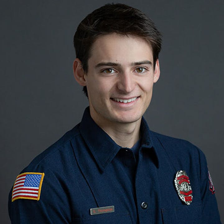 A person in a dark uniform with an American flag patch and badge, smiling against a plain background.