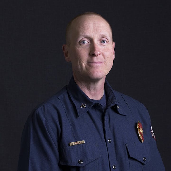 A person in a navy blue uniform with a badge poses for a portrait in front of a dark background.