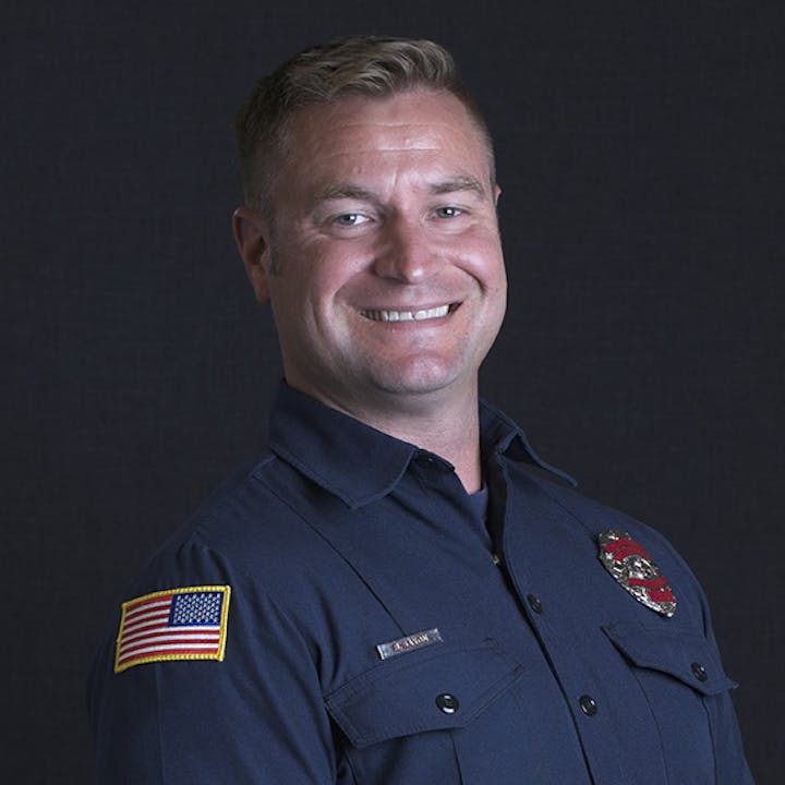 A smiling person in a blue uniform with an American flag patch and a badge.