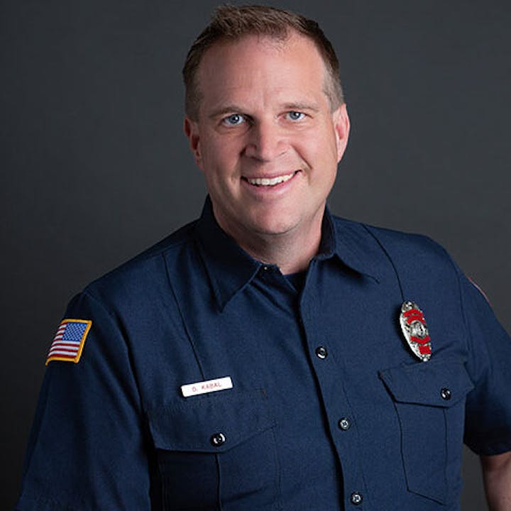 A person in a dark uniform with badges and an American flag patch, smiling against a plain background.