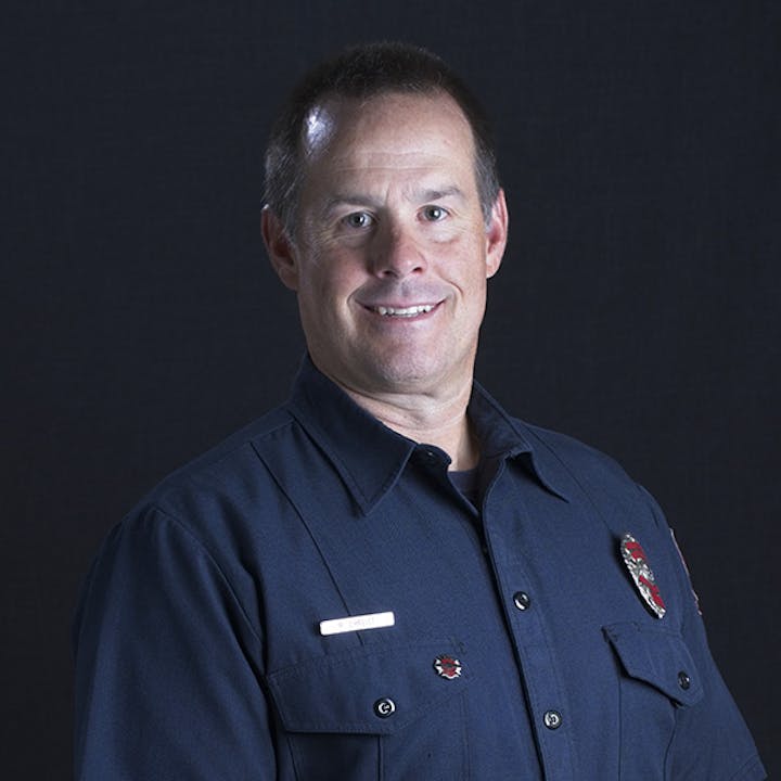 A person in a dark uniform with badges, smiling against a plain background.