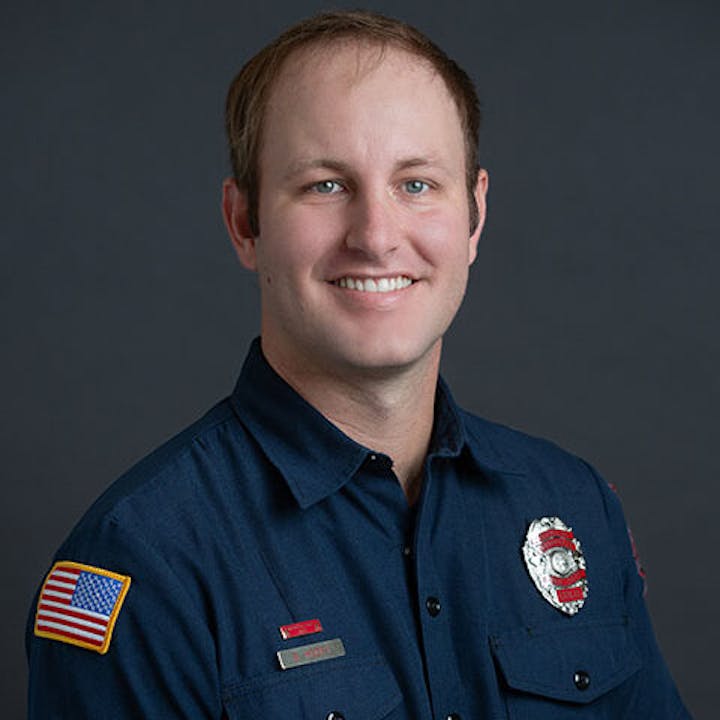 A person in a blue uniform with a firefighter badge and American flag patch, smiling against a dark background.