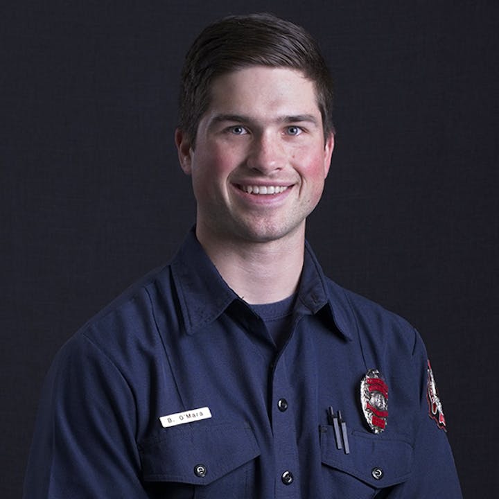 Smiling man in a dark uniform with name tag and badge against a dark background.
