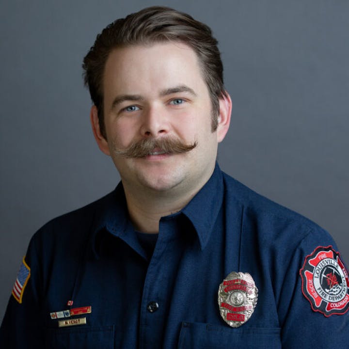 A man in a blue uniform with a mustache, wearing a badge and patch, posing for a portrait against a gray background.