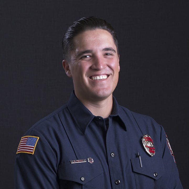 A smiling person in a dark uniform with a U.S. flag patch and a badge, against a dark background.