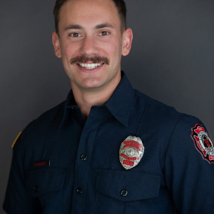 A person in a dark uniform with a badge and patches, smiling in a professional portrait.