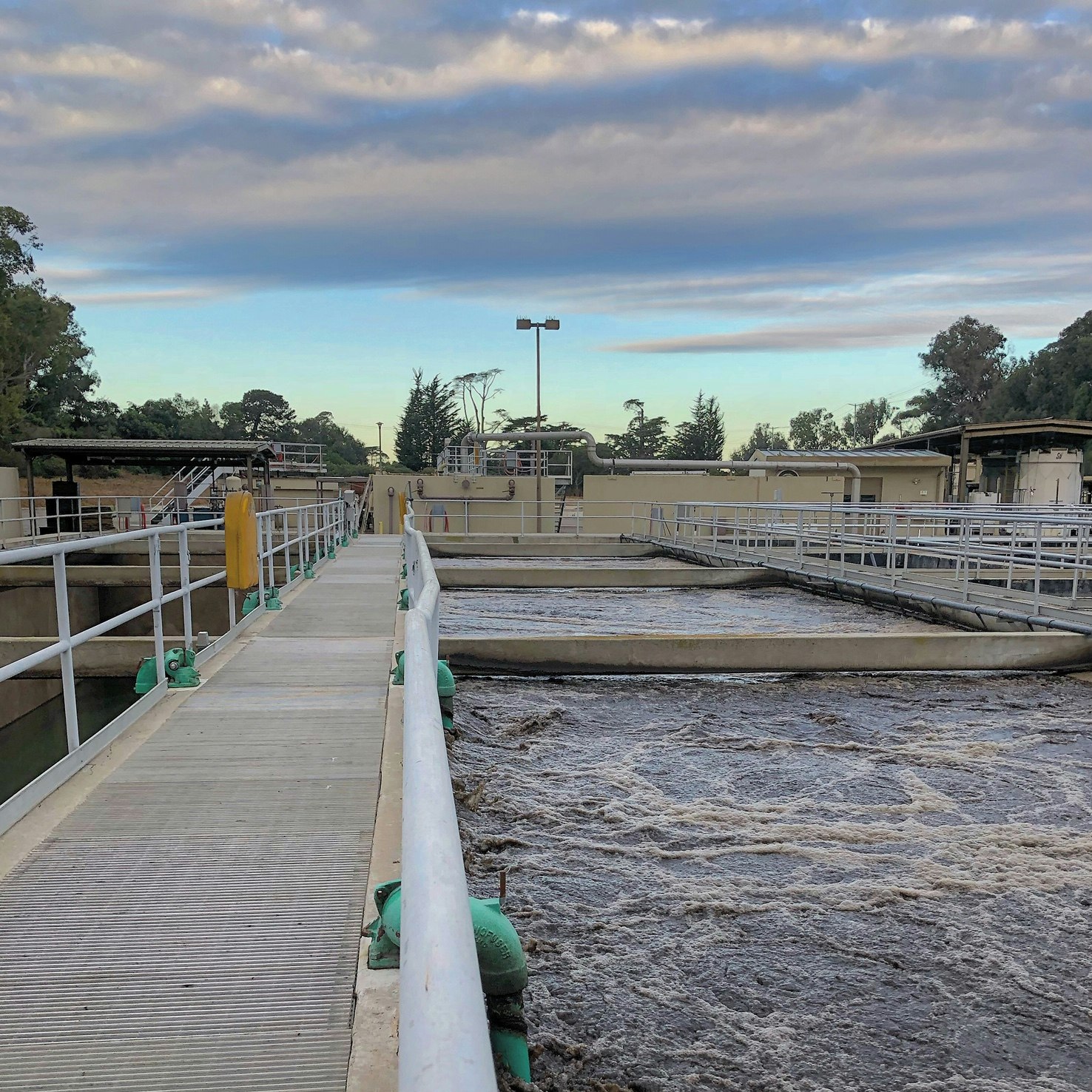 The image shows a water treatment facility with open tanks and walkways, set against a cloudy sky.