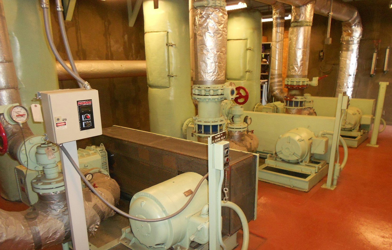 Industrial room with machinery, pipes, and control panels on a red floor.