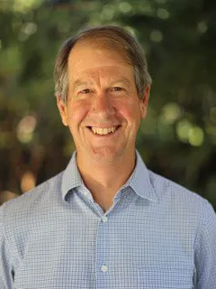 The image shows a man with a friendly smile, wearing a light blue checked shirt, set against a blurred green background.