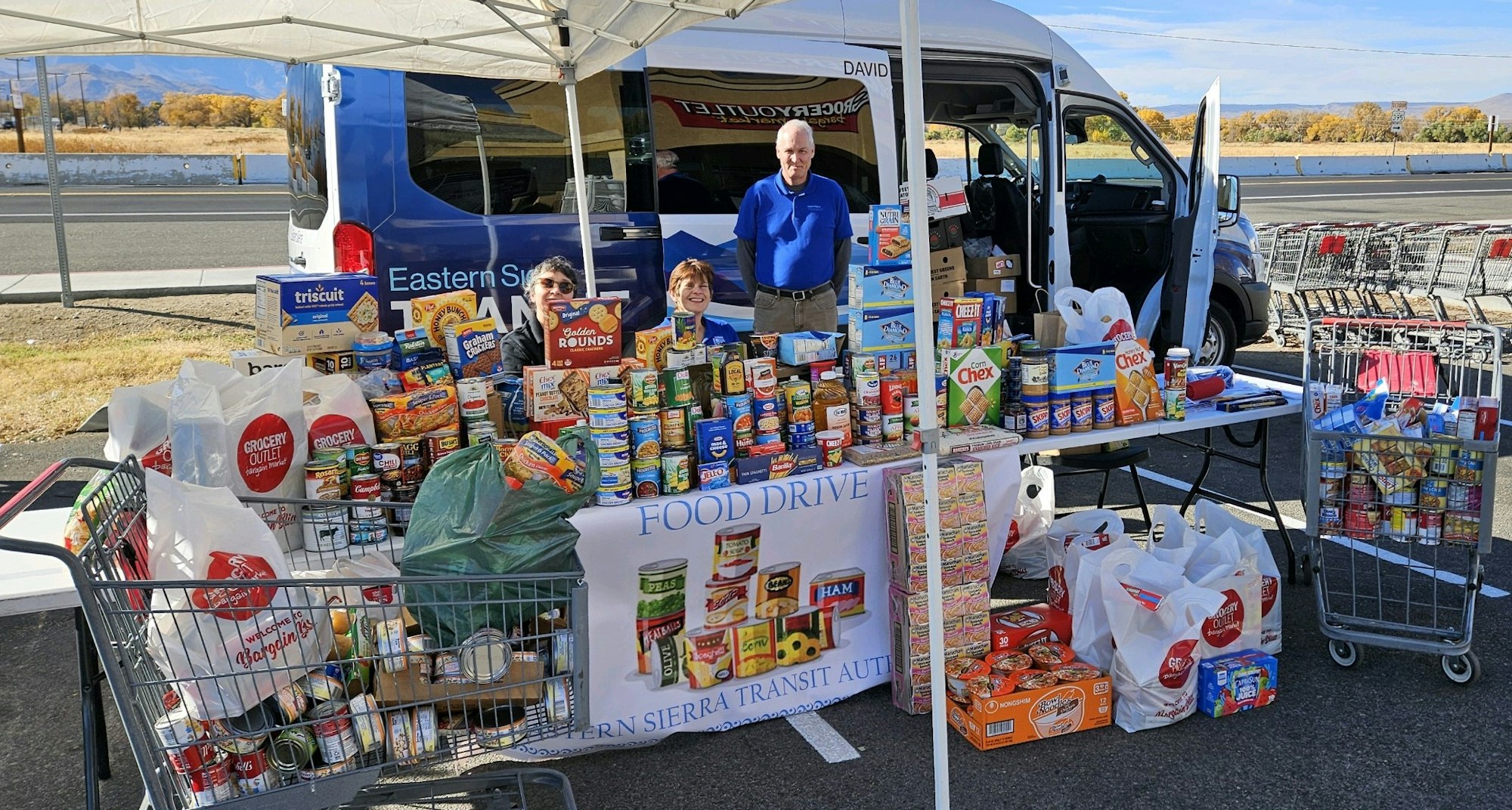 A food drive with people, non-perishable foods, shopping carts, and a van under a tent.