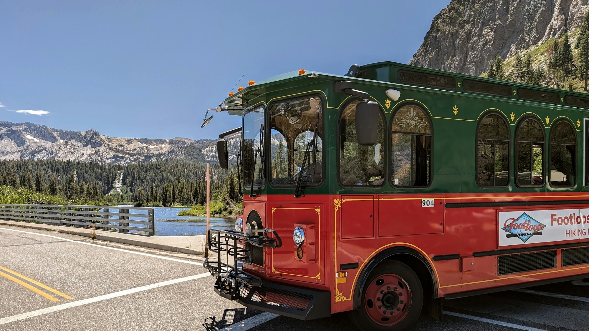 A red and green trolley bus on a road with a mountainous backdrop and a lake.
