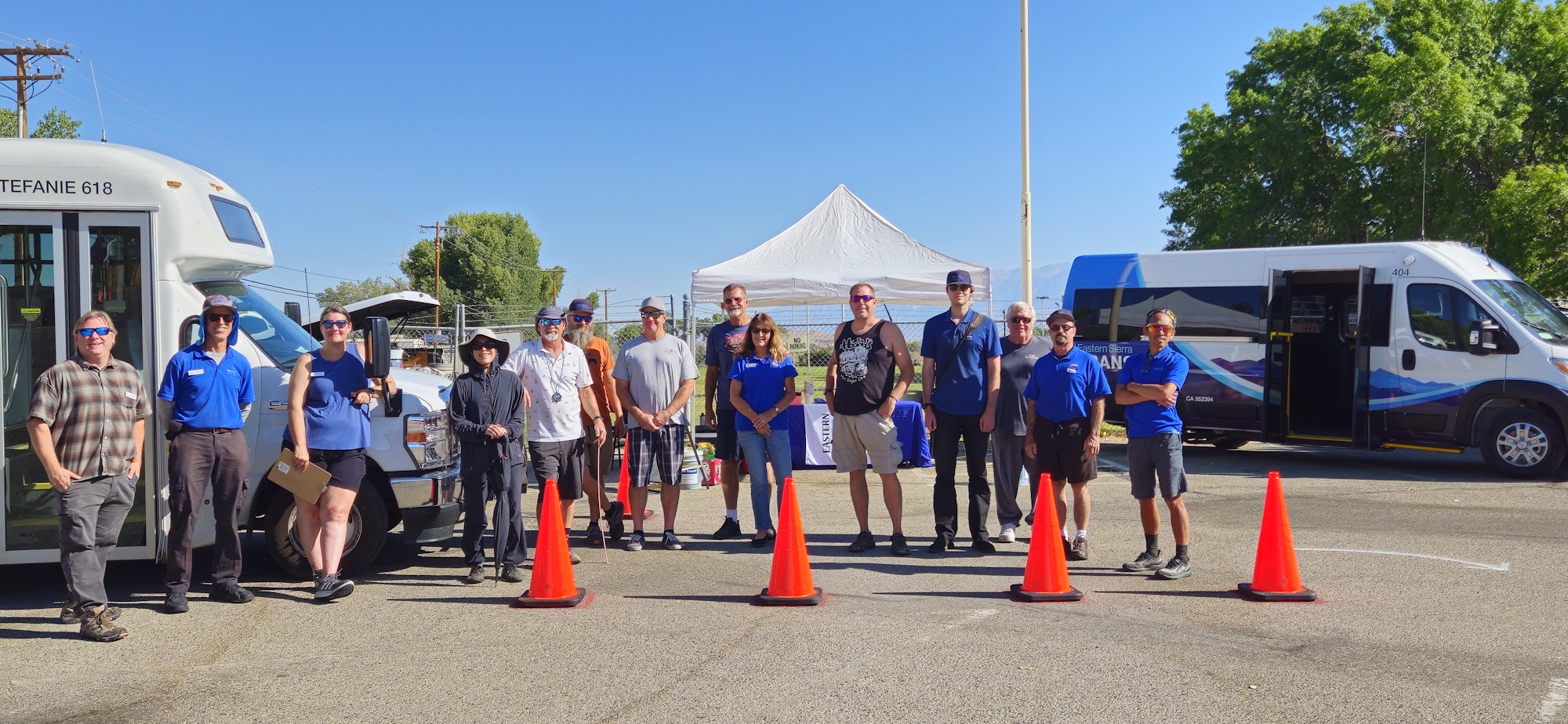 A group of people stands between two transit vans, with traffic cones and a white canopy in the background on a sunny day.
