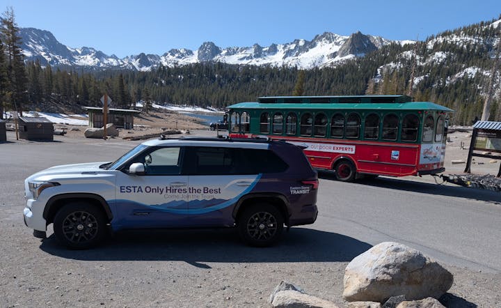 Mountainous landscape with a transit SUV and a red trolley bus on a paved road, surrounded by snowy peaks and pine trees.