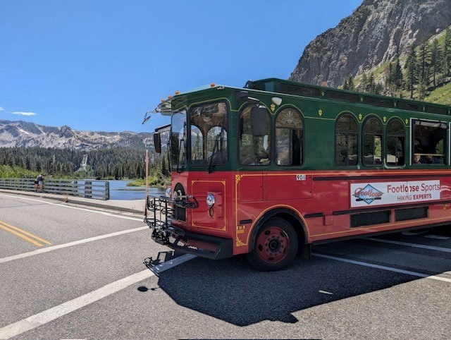 A red trolley-style bus parked on a road near a lake with mountains in the background and a clear blue sky.