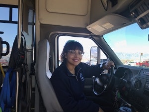 A smiling person in a vehicle, possibly a truck or large equipment, with a steering wheel and windows showing a bright day outside.