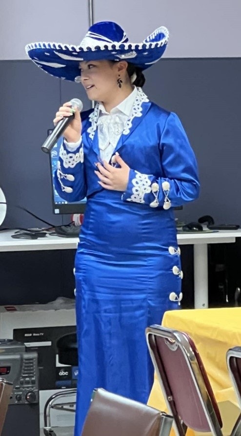 A person in a blue traditional outfit and hat is speaking into a microphone in a room with chairs and a table with a yellow cloth.